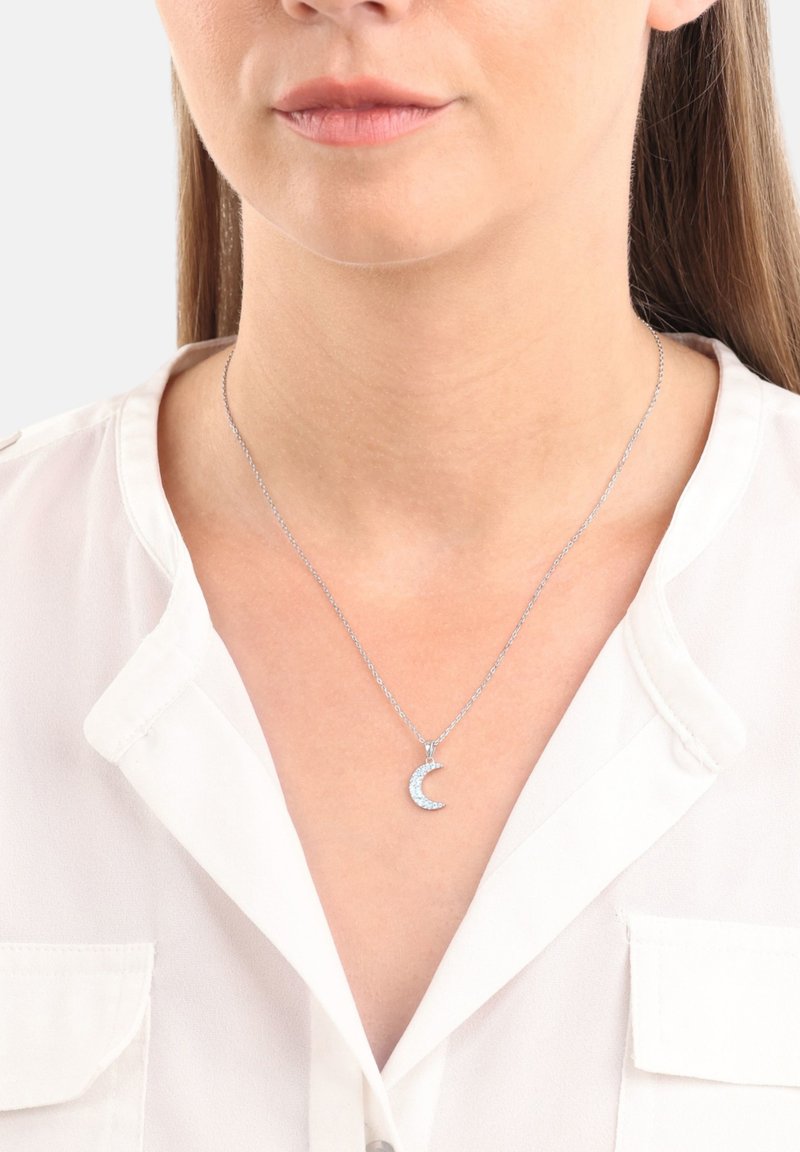 Collana d'argento con pendente a forma di mezzaluna, impreziosita da piccole pietre. La catena è delicata, in perfetto abbinamento a una blusa bianca.