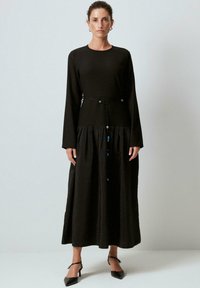 Musta pitkähiha dressi, jossa on jämäkkä yläosa, poimutettu hame ja kiristettävä vyötärö. Sisältää koristeellisia metalliosia.