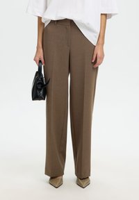 Femme portant un pantalon droit marron, des chaussures beiges à bout pointu, un haut blanc surdimensionné, tenant un sac à main noir avec un détail noué.
