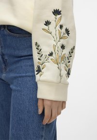 Sudadera crema con bordado floral en verde y azul marino en la manga, combinada con jeans de mezclilla azules. Textura suave y corte relajado.