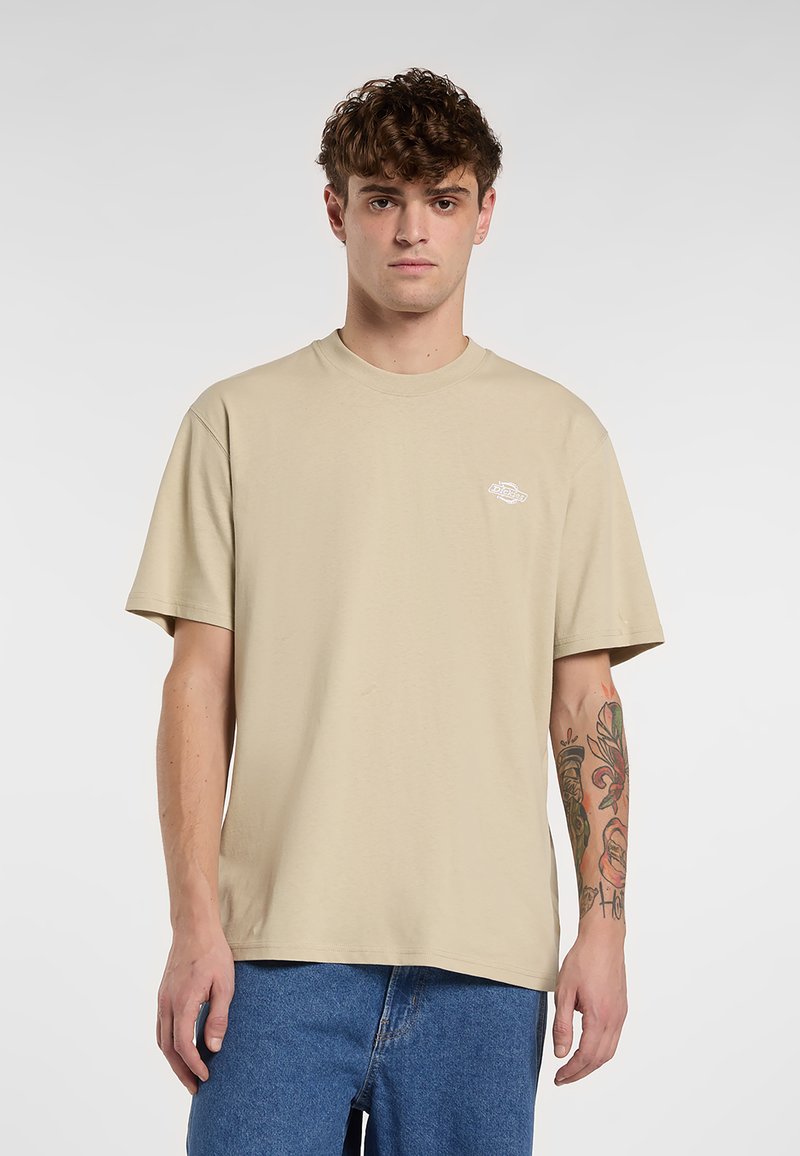 Beige katoenen T-shirt met een ronde hals en korte mouwen, voorzien van een klein, wit logo op de linkerborst. Draagt bij een blauwe spijkerbroek.