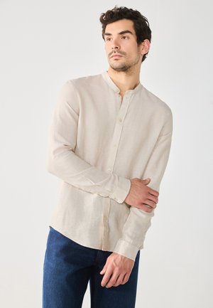 Light beige, long-sleeve, button-up shirt with a mandarin collar and subtle texture. Paired with dark blue jeans.