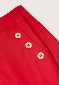 Vêtement en tissu rouge doté de trois boutons dorés à l'avant. Le matériau est lisse avec une finition nette et ajustée.