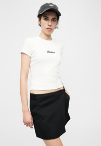 Νεαρό άτομο φορώντας λευκό εφαρμοστό T-shirt με το λογότυπο «Dickies», μαύρη φούστα και σκούφο μπέιζμπολ σκούρο, στέκεται μπροστά σε απλό φόντο.