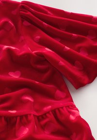 Tessuto di velluto morbido rosso con motivi di piccoli cuori rosa. Il design presenta maniche a sbuffo e una vita arricciata.