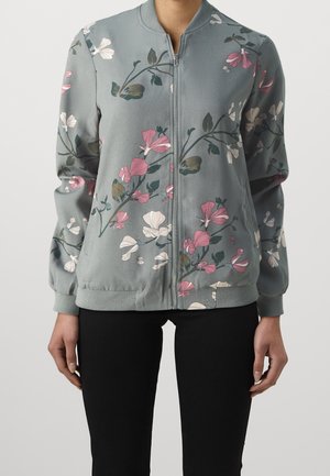 Jasnoszara bomberka na suwak z różowymi, białymi i zielonymi kwiatowymi gałązkami, noszona z czarnymi spodniami przez osobę stojącą na białym tle.