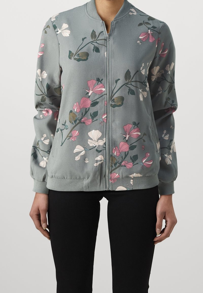 Jasnoszara bomberka na suwak z różowymi, białymi i zielonymi kwiatowymi gałązkami, noszona z czarnymi spodniami przez osobę stojącą na białym tle.