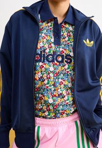 Marinblå zip-jacka med gula ränder, blommig polo med flerfärgade mönster och rosa shorts med gröna sidostripes.