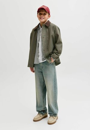 Garçon souriant portant une casquette rouge, une veste verte, une chemise à carreaux, un t-shirt blanc, un jean bleu ample et des chaussures beiges, debout avec une main dans la poche.