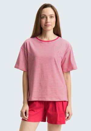 Frau mit langen braunen Haaren, die ein rosa-weiß gestreiftes kurzärmeliges Oberteil und einfarbige rosa Shorts trägt, steht vor einem schlichten Hintergrund.