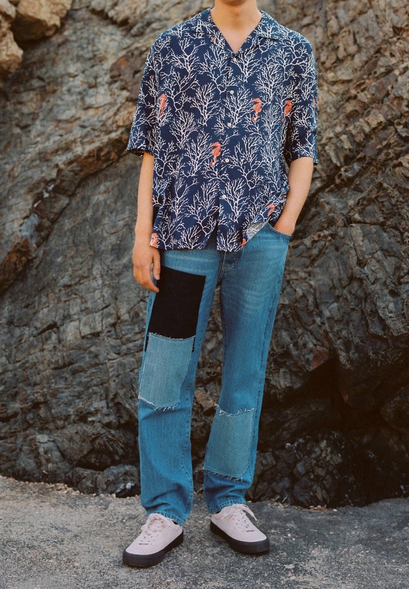 Personne portant une chemise bleu marine avec des coraux blancs et des hippocampes orange, un jean patchwork et des baskets rose clair debout près d'une surface rocheuse.