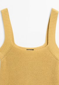 Débardeur beige en tricot avec larges bretelles et encolure carrée, montrant une texture détaillée et une petite étiquette de marque noire à l’intérieur.