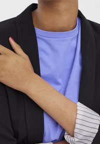 Černý blazer přes světle modré tričko, s bílou pruhovanou košilí, která vykukuje z rukávu blazera. Dobře viditelná oblast kolem paží a krku.