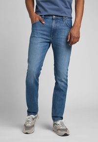 Niebieskie dżinsy o slim fit, pięciokieszeniowym kroju i delikatnym wyblaknięciu. Połączone z jasnoszarymi trampkami i niebieską koszulą.