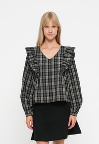 Blouse à carreaux noir et blanc avec épaules volantées, encolure en V et manches longues, portée avec une jupe noire ajustée. Tissu lisse et léger.