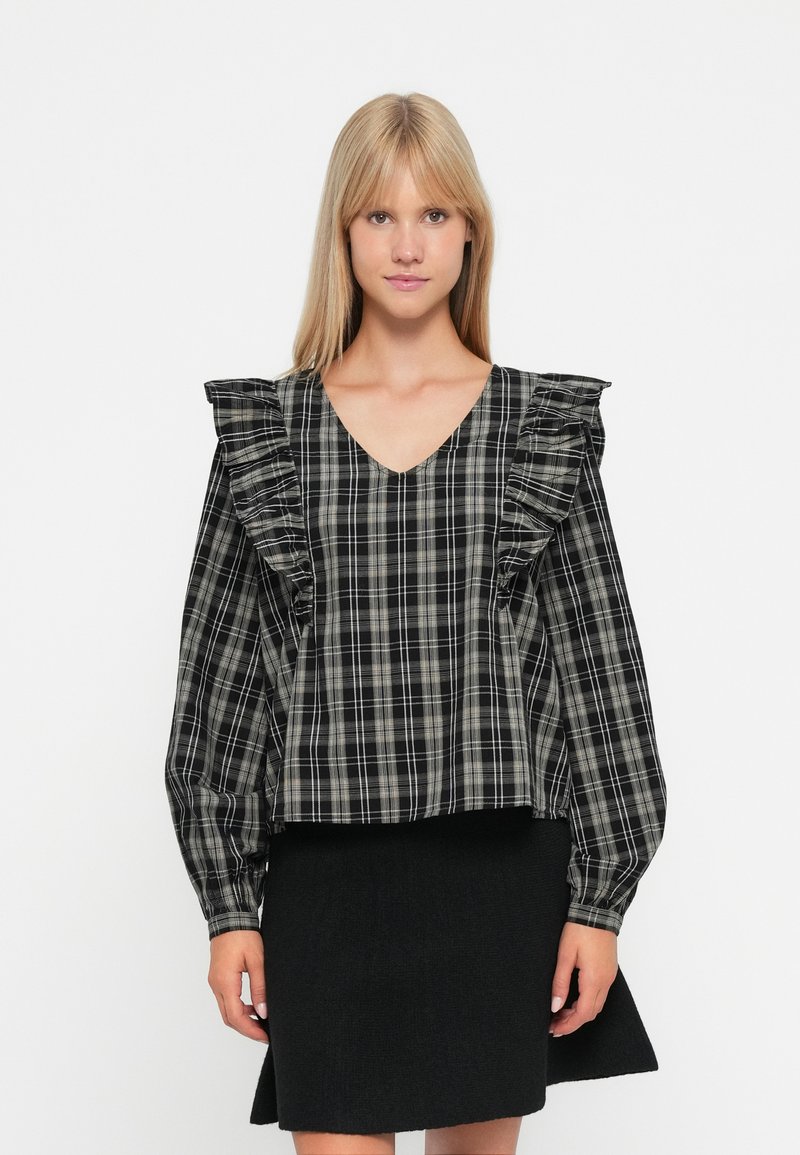 Blouse à carreaux noir et blanc avec épaules volantées, encolure en V et manches longues, portée avec une jupe noire ajustée. Tissu lisse et léger.