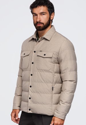 Homme aux cheveux foncés et à la barbe porte une veste matelassée beige avec des poches poitrine boutonnées et des boutons noirs, debout devant un fond clair.