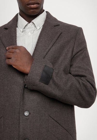 Hnedý kabát z vlnenej zmesi s elegantným dizajnom, ktorý má čierny kožený detail na manžetách a zapínanie na gombíky s dvoma bočnými vreckami.