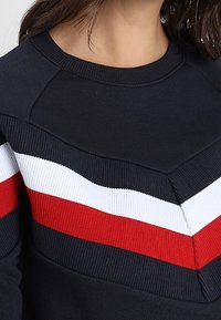 Felpa blu navy con finitura testurizzata, caratterizzata da un motivo a chevron con strisce rosse, bianche e blu navy sulla zona del petto e delle spalle.
