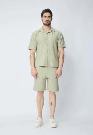 Homme à cheveux courts et foncés avec des tatouages, portant une chemise à manches courtes à motifs vert clair et un short assorti, avec des baskets blanches.