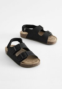 Svarta sandaler med två justerbara remmar med spännen, en korkfotbädd och en texturerad mockainläggssula. Öppen design för ventilering.