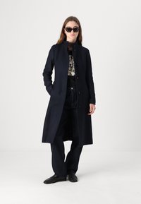 Marinblå ull lång kappa med hög krage, knäppning fram och sidofickor, tillsammans med en mönstrad topp och mörka denimbyxor.