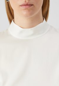 Nahbilde er en person som har på seg en hvit topp med høy hals, og bildet viser nakken, nedre del av ansiktet og øvre del av brystet mot en nøytral bakgrunn.