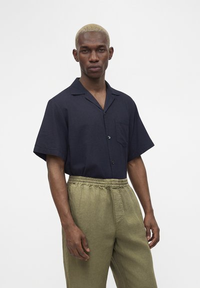 Άνδρας με κοντά ξανθά μαλλιά, φοράει ένα σκούρο κοντομάνικο πουκάμισο με κουμπιά, κεντημένο σε ελαστικό παντελόνι χακί, στέκεται μπροστά σε λευκό φόντο.