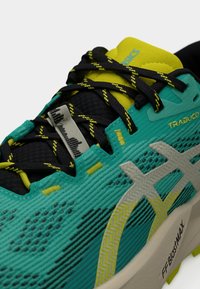 Αθλητικό παπούτσι σε τιρκουάζ και μαύρο με υφή στο επάνω μέρος, κίτρινες λεπτομέρειες και περίπλοκο δέσιμο κορδονιών. Διαθέτει χοντρή σόλα και αντανακλαστικές λεπτομέρειες.