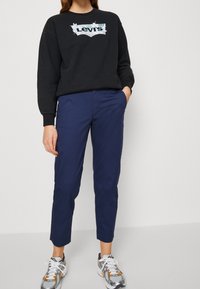 Sweatshirt noir avec un logo bleu Levi's sur la poitrine, associé à un pantalon slim-fit bleu marine et des baskets grises avec des accents orange.