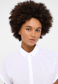 Camisa blanca de botones hecha de una tela liviana, con un corte holgado, mangas cortas y un logo sutil en el lado izquierdo del pecho.