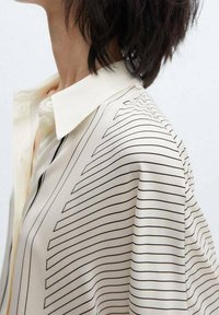 Beige Seidenhemd mit schwarz gestreiftem Muster. Das Hemd hat einen spitzen Kragen und ein entspanntes, fließendes Design.