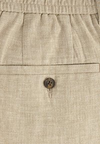 Tissu beige texturé avec une poche à rabat horizontale et un bouton marron foncé à quatre trous sous une ceinture froncée.