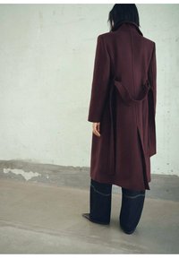 Manteau en laine bordeaux au design structuré, à manches longues et avec un détail à la taille nouée, associé à un jean large noir et des chaussures pointues.