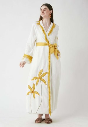 White long dress with a yellow tie belt, featuring yellow embroidered leaves and fringed accents on the collar and sleeves. Brown sandals.