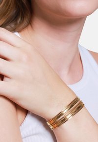 Goldene Armreifen an einem hellen Handgelenk, die eine glatte, glänzende Oberfläche mit unterschiedlichen Breiten und strukturierten Akzenten aufweisen.