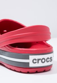 Crvene gumene Crocs sandale s prorezanom trakom, koje imaju debeli đon s crno-bijelim prugama i istaknutim "crocs" logotipom sa strane.