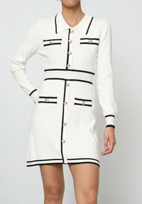 Robe tricotée blanche à manches longues, avec des accents noirs et un col. Présente des boutons sur le devant et deux poches poitrine.