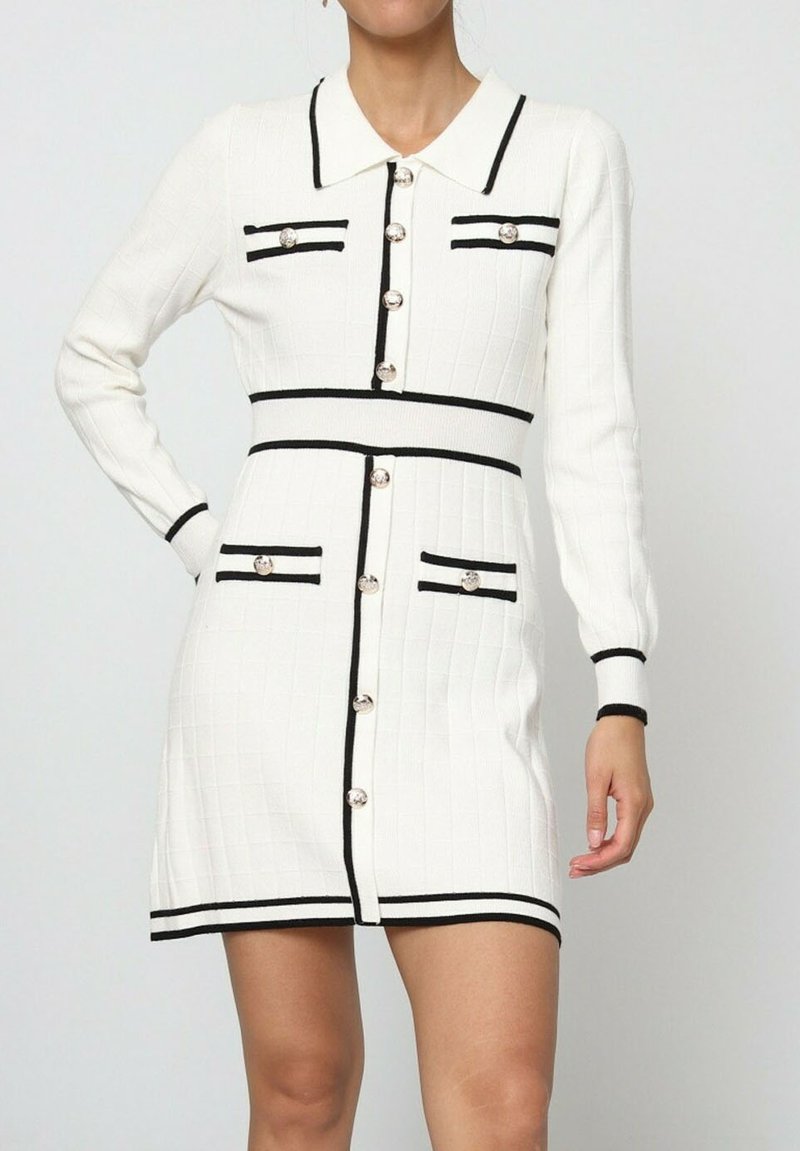 Robe tricotée blanche à manches longues, avec des accents noirs et un col. Présente des boutons sur le devant et deux poches poitrine.