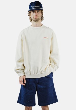 Hombre con sudadera color crema con texto "Pockies", pantalones cortos azul marino y gorra azul marino con texto "Weekend", de pie con una mano en el bolsillo.