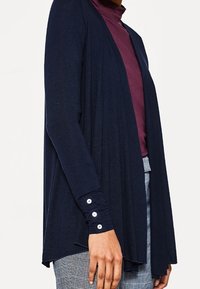 Marineblauer, leichter Cardigan mit offenem Vorderteil und langen Ärmeln, der Knopfdetails an den Bündchen aufweist. Texturiertes, drapiertes Design.