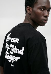 Jeune homme regardant par-dessus son épaule, portant un t-shirt noir avec un texte blanc « Dream Big, Grind Bigger » dans le dos sur fond uni.