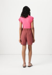 Haut rose ajusté à manches courtes avec volants, associé à un short ample de couleur rose, arrivant à mi-cuisse. Le modèle porte des sandales plates.