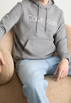 Person trägt einen grauen Calvin Klein Hoodie und hellblaue Jeans und sitzt auf einem strukturierten beigen Stuhl.