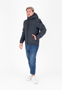 Giacca da uomo grigia scura trapuntata con cappuccio, dotata di una chiusura lampo frontale, due tasche laterali e una finitura tessurizzata. Abbinata a jeans blu e sneaker bianche.