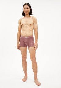 Jeune homme avec des cheveux foncés de longueur moyenne, souriant, portant des boxer mauves, debout pieds nus sur un fond blanc uni.