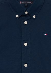 Camisa azul-marinho de botão, feita de algodão, com colarinho clássico, botões em marfim e um pequeno logótipo bordado em vermelho, branco e azul.