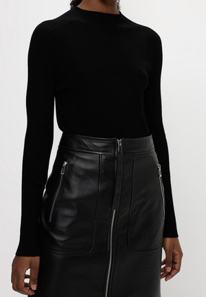 Femme portant un haut côtelé noir à manches longues et une jupe en cuir noire avec fermeture éclair devant et deux poches zippées.