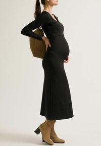 Schwarzes, langärmliges Umstandskleid mit einem gewellten Ausschnitt, kombiniert mit braunen Wildleder-Ankle-Boots und einer braunen Handtasche. Schlichte, elegante Silhouette.