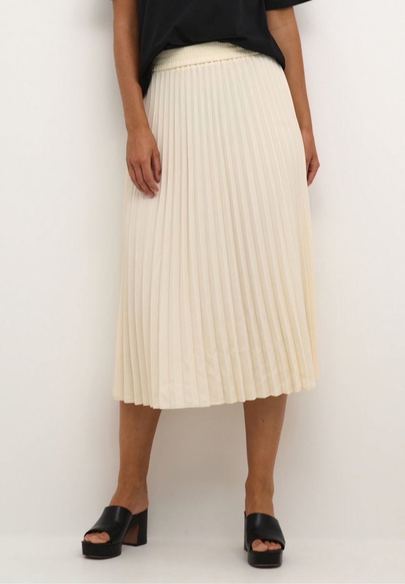 Falda midi plisada en tejido crema, con superficie texturizada y cintura elástica. Combinada con sandalias negras de plataforma gruesa.